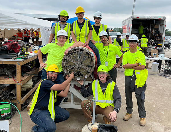 A group of people in safety vests and hard hats standing around a large cylindrical piece of machinery at an outdoor worksite. The background includes tools, equipment, a workbench, a tent, and a truck. The scene suggests a team effort or project, with the group posing for a photo.