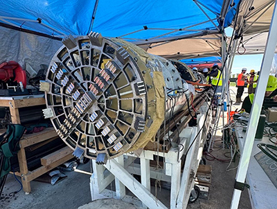 Eine große zylindrische Bohrmaschine, die für den Tunnel- und Aushubbau eingesetzt wird und auf einem weißen Holzständer unter einem blauen Vordach positioniert ist. Die Maschine hat an einem Ende eine gitterartige Struktur. Im Hintergrund sind mehrere Personen in Warnwesten und Helmen zu sehen, die auf eine Baustelle im Freien oder einen Testbereich hindeuten.