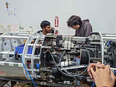 Two individuals working on a large cylindrical boring machine, which is partially assembled with motors, wiring, and structural elements. One person holds a tool, while the other assists.