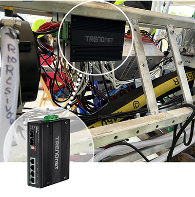 A TRENDnet TI-PG62B industrial switch installed within a complex setup of wires and equipment. The switch is highlighted with a circular zoom-in, showing its ports and connectors. It is mounted on a metal frame, surrounded by various cables and components, indicating its use in an industrial environment. The text "TRENDnet" is visible on the switch