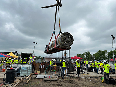 A large cylindrical boring machine being lifted by a crane using straps and cables at a busy construction site. Numerous workers in yellow safety vests and hard hats are present, some observing and others actively working. The site is filled with various equipment and materials, including pallets, containers, and tools. The sky is overcast, with tents and trees visible in the background.